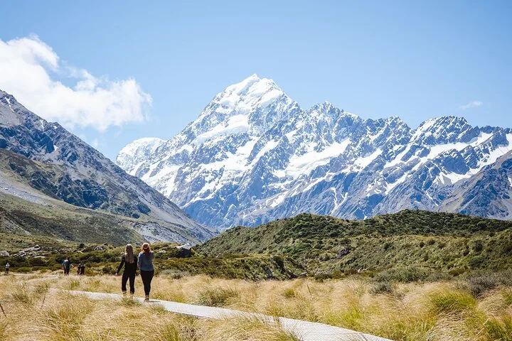Two walkers on the Hooker Valley Track path through golden tussock toward Aoraki/Mount Cook